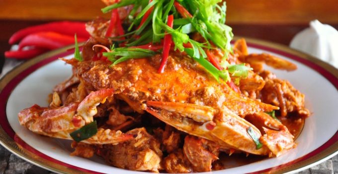 A close-up image of a plate of Chili Crab, showcasing whole mud crab pieces smothered in a rich, tangy chili sauce, garnished with fresh cilantro leaves and served with steamed buns on the side.